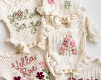 May include: Five cream-colored ribbed knit rompers featuring long sleeves, ruffled leg openings, and wooden buttons. Each garment is personalized with embroidered names including Saylor, Shiloh Sage, Vella Rue, and Gracie, alongside floral accents.