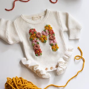 May include: Cream-colored baby romper with long sleeves and a ruffled bottom. The letter "M" is embroidered on the front with colorful flowers in shades of pink, orange, and yellow. Wooden buttons fasten the romper.