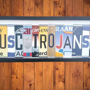 May include: A framed wall art piece made from vintage license plates spelling out "USC TROJANS". The frame is black and the license plates are a variety of colors, including red, white, blue, and yellow.