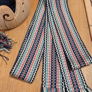 May include: A woven textile with a striped pattern in navy, white, teal, and coral. The textile has fringed ends and is displayed on a wooden surface. Yarn and a wooden yarn bowl are visible in the background.