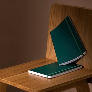 Puede incluir: Dos cuadernos de color verde oscuro con cierres elásticos están apilados en una silla de madera. Los cuadernos están cerrados y tienen un diseño sencillo.