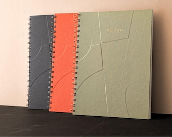 May include: Three spiral-bound notebooks in dark gray, orange, and sage green. Each cover features a debossed geometric line pattern and the text HOXON printed in gold. The notebooks are arranged in a row, showing their front covers and metal coils.