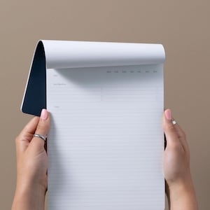 May include: A white notepad with a dark blue backing, featuring a printed layout with sections for date, key objectives, and notes, including days of the week from Monday to Sunday and four circular checkboxes for task tracking on each line.
