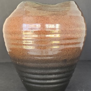 May include: A handcrafted ceramic vase with a unique, organic shape. The vase features a gradient of colors, transitioning from a dark black base to shades of brown and tan at the top. The textured surface and ribbed lower section add visual interest.