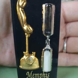 May include: A black souvenir featuring a gold telephone and a silver hourglass design. The word "Memphis" is written in gold script at the bottom. The items are likely a magnet or a decorative piece.