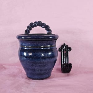May include: A deep blue ceramic wall pocket with a glossy, speckled finish and a twisted rope-style handle at the top. Beside the ceramic vessel is a black metal wall hook featuring a vertical mounting plate with a single screw hole at the top.