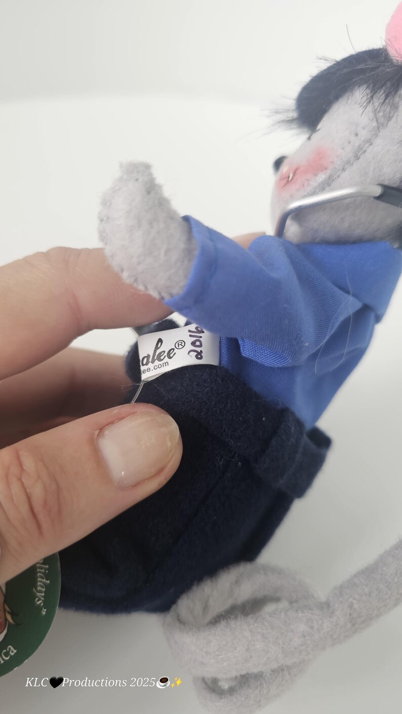 Puede incluir: Un rat&oacute;n de peluche gris con camisa azul, pantalones azul marino y gafas. El rat&oacute;n tiene la nariz y las mejillas rosas. Una etiqueta dice "alee&reg; ee.com" y "2016". El rat&oacute;n es un juguete de peluche con tem&aacute;tica navide&ntilde;a.