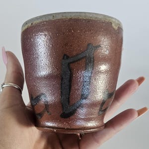May include: A handmade, brown ceramic cup with a textured surface. The cup features a dark, abstract design painted on the exterior. The rim has a lighter, tan color. The cup is held in a hand.