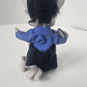 Puede incluir: Un mu&ntilde;eco de rat&oacute;n de peluche con una camisa azul, una falda negra y un sombrero azul oscuro. El rat&oacute;n tiene orejas y patas grises. La mu&ntilde;eca se ve desde atr&aacute;s, con los brazos extendidos. El fondo es blanco.