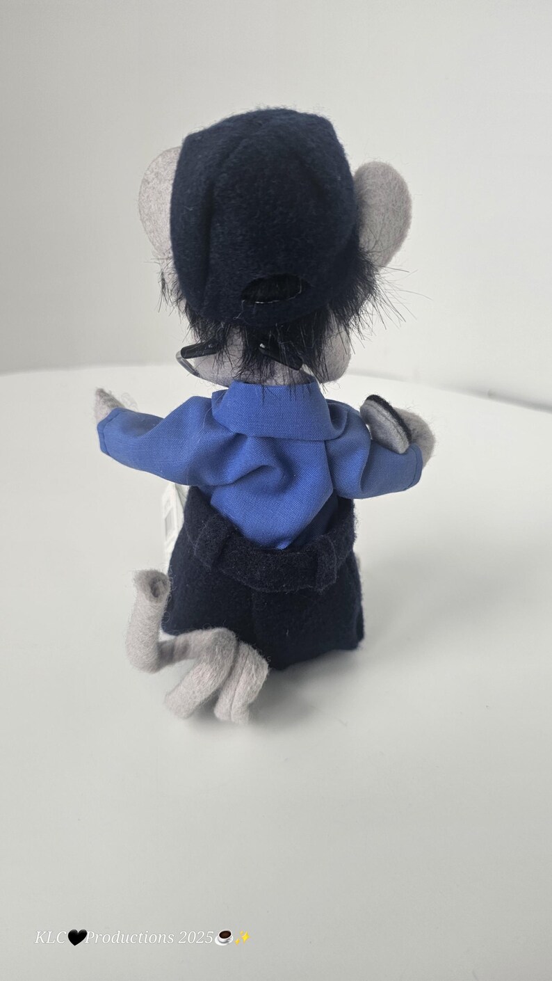 Puede incluir: Un mu&ntilde;eco de rat&oacute;n de peluche con una camisa azul, una falda negra y un sombrero azul oscuro. El rat&oacute;n tiene orejas y patas grises. La mu&ntilde;eca se ve desde atr&aacute;s, con los brazos extendidos. El fondo es blanco.