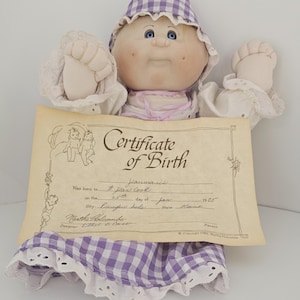 May include: A vintage doll with a round face, blue eyes, and a purple and white checkered bonnet and dress. The doll holds a "Certificate of Birth" with handwritten details. The doll's arms and hands are visible.