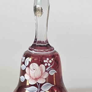 May include: A clear glass bell with a burgundy-colored base and handle. The bell is decorated with a hand-painted pink rose and white leaves. A small, round, gold-colored sticker is affixed to the handle. The bell is likely a decorative item.