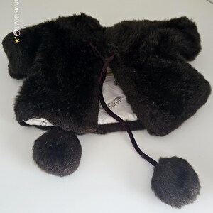May include: A dark brown faux fur bolero jacket with a tie closure and two pom-poms. The jacket has a soft, plush texture and a white lining. The bolero is designed to be worn over other garments.