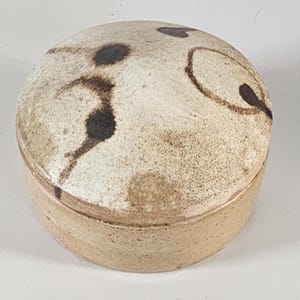 May include: A round, ceramic trinket box with a lid. The box is beige with dark brown abstract designs on the lid. The box has a simple, minimalist design.