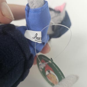 Puede incluir: Un juguete de peluche de gato gris con un atuendo azul. El juguete tiene una nariz rosa y una etiqueta que dice "Anna". El gato sostiene una etiqueta que dice "Home for the Holidays".