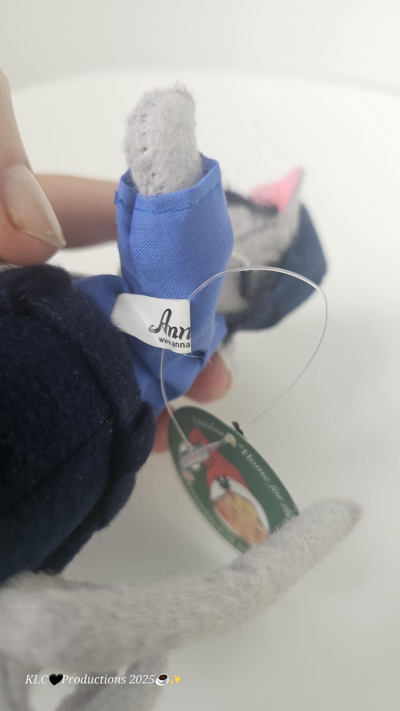 Puede incluir: Un juguete de peluche de gato gris con un atuendo azul. El juguete tiene una nariz rosa y una etiqueta que dice "Anna". El gato sostiene una etiqueta que dice "Home for the Holidays".