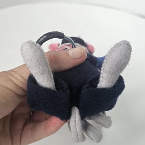 Puede incluir: Un mono de peluche con brazos y pies grises, cuerpo azul marino y un anillo negro. El mono tiene una cara rosa y est&aacute; sostenido por una mano.