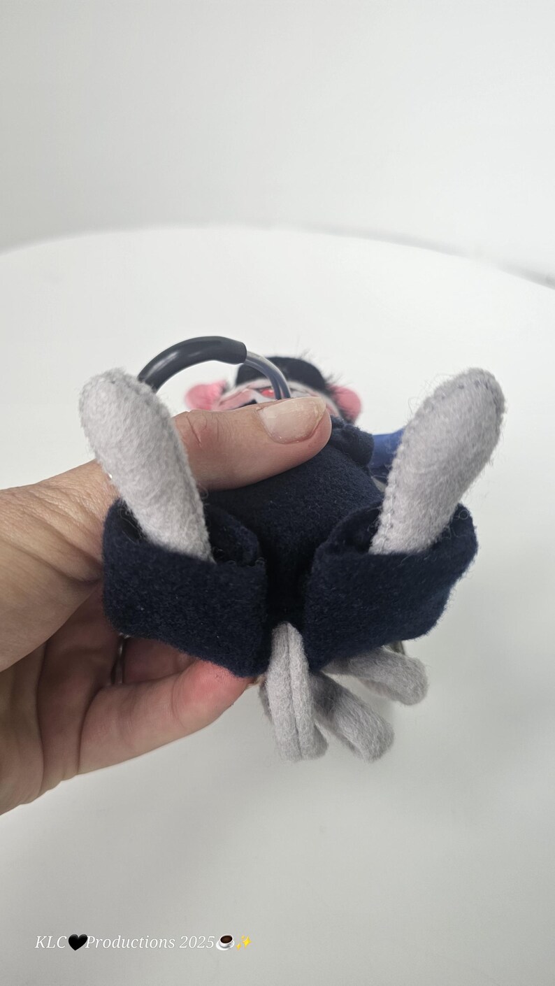 Puede incluir: Un mono de peluche con brazos y pies grises, cuerpo azul marino y un anillo negro. El mono tiene una cara rosa y est&aacute; sostenido por una mano.