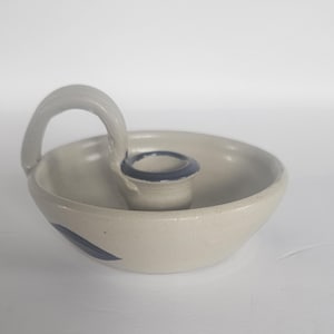 May include: A ceramic candle holder with a light gray finish and a blue accent. The candle holder has a circular bowl shape with a handle and a central candle cup. A blue leaf design is painted on the side.