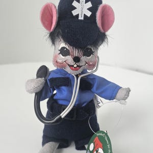Puede incluir: Una figura de rat&oacute;n de peluche vestida de param&eacute;dico, con uniforme azul marino y gorra con un s&iacute;mbolo m&eacute;dico blanco. El rat&oacute;n tiene orejas rosas y sostiene un estetoscopio. Una etiqueta dice "Home for the Holidays".