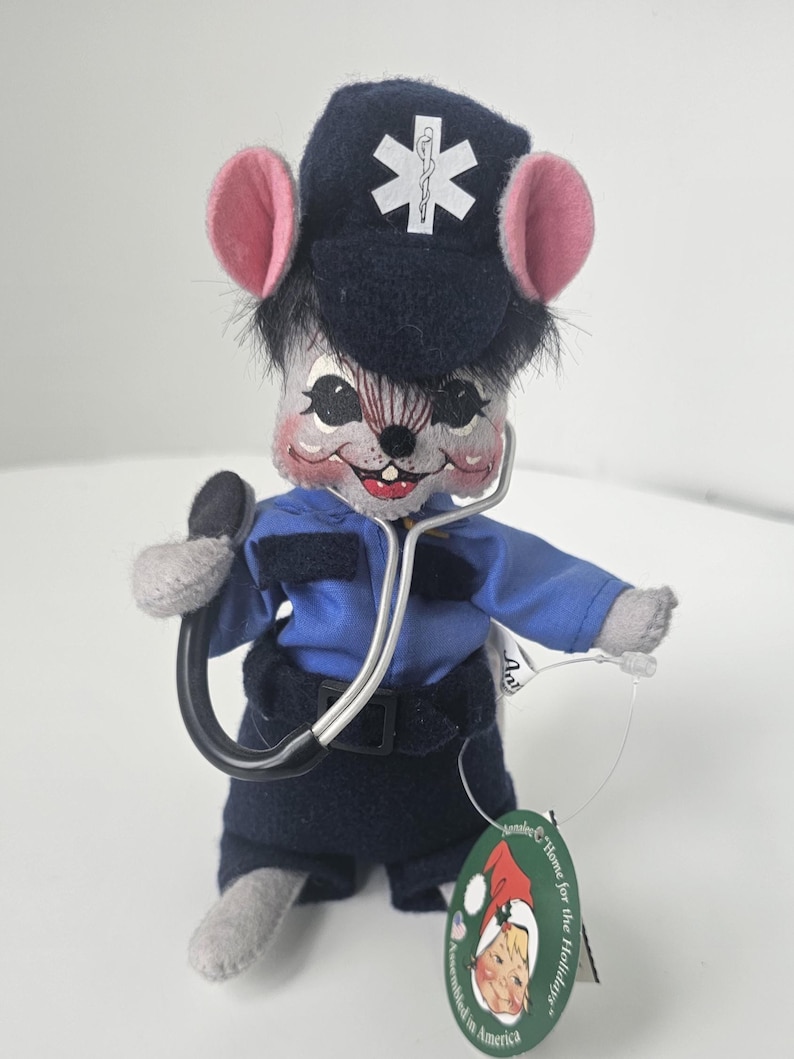 Puede incluir: Una figura de rat&oacute;n de peluche vestida de param&eacute;dico, con uniforme azul marino y gorra con un s&iacute;mbolo m&eacute;dico blanco. El rat&oacute;n tiene orejas rosas y sostiene un estetoscopio. Una etiqueta dice "Home for the Holidays".