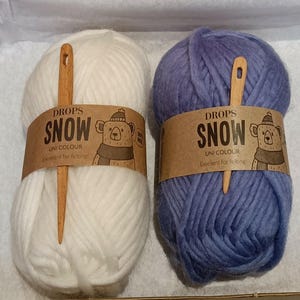 Op de afbeelding: Twee bollen Drops Snow garen, één wit en één blauw, elk omwikkeld met een bruin papieren label met een berenillustratie en de tekst Drops Snow Uni Colour. Een houten naald is verticaal in het midden van elke bol garen gestoken.
