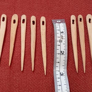 May include: A selection of wooden yarn needles, of varying sizes, are arranged on a red surface. A measuring tape is placed next to the needles, showing measurements in centimetres. The needles are light brown and have a hole at the top.