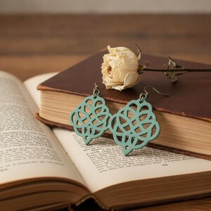 May include: A pair of sage green dangle earrings featuring an intricate, symmetrical, openwork knot design. The earrings are suspended from metal hooks and are displayed alongside a dried cream-colored rose on a closed, dark-bound vintage book.