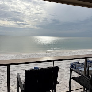 Op de afbeelding: Een balkonuitzicht met twee zwarte rieten stoelen met metalen frames achter een houten reling. Het tafereel kijkt uit over een wit zandstrand en een kalme oceaan onder een bewolkte lucht, waarbij de zon reflecteert op het wateroppervlak.