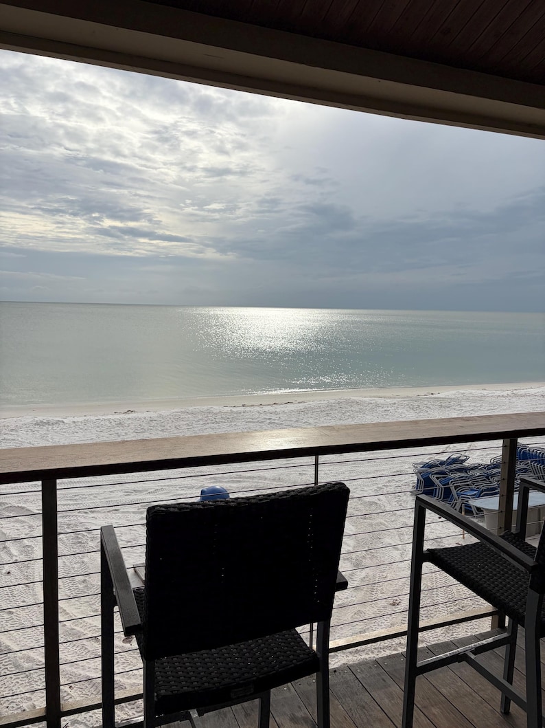 Op de afbeelding: Een balkonuitzicht met twee zwarte rieten stoelen met metalen frames achter een houten reling. Het tafereel kijkt uit over een wit zandstrand en een kalme oceaan onder een bewolkte lucht, waarbij de zon reflecteert op het wateroppervlak.