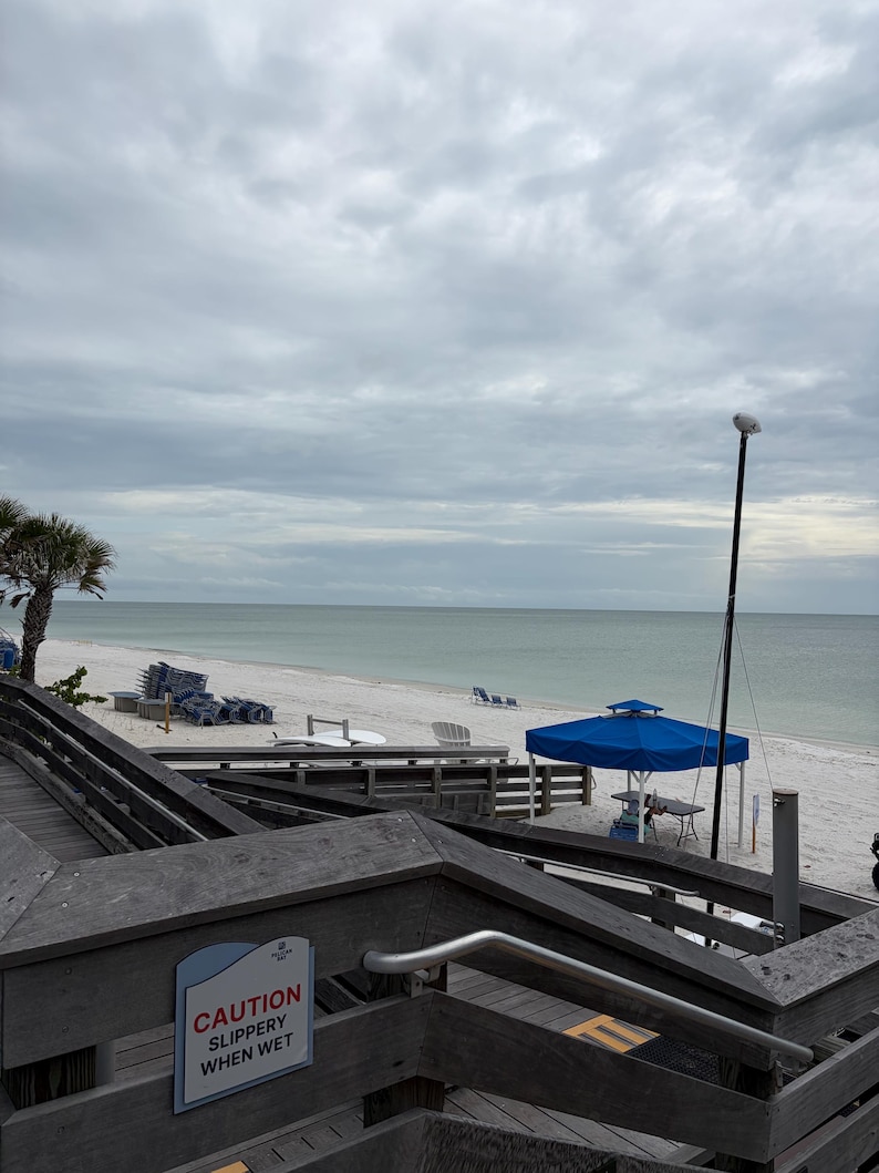 Op de afbeelding: Een houten trap die naar een zandstrand leidt onder een bewolkte lucht. Een bord op de reling vermeldt CAUTION SLIPPERY WHEN WET. Het strand is voorzien van een blauwe overkapping, verspreide stoelen en een kalme oceaanhorizon onder een grijze lucht.