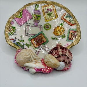 Op de afbeelding: Een decoratieve oesterschelpschaal met een goudkleurige rand, voorzien van een decoupagepatroon met kleurrijke illustraties van bloemen, vazen en rechthoekige kaders. De basis is versierd met drie kleinere schelpen en roze kralen.