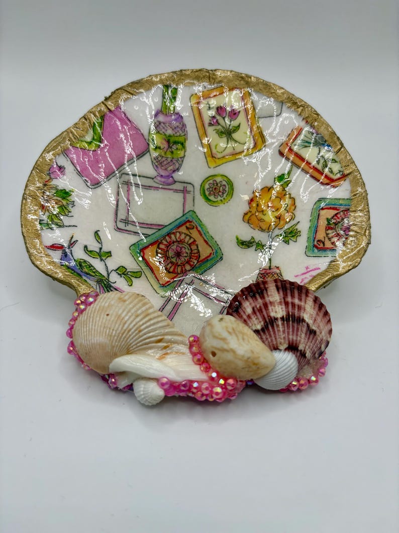 Op de afbeelding: Een decoratieve oesterschelpschaal met een goudkleurige rand, voorzien van een decoupagepatroon met kleurrijke illustraties van bloemen, vazen en rechthoekige kaders. De basis is versierd met drie kleinere schelpen en roze kralen.