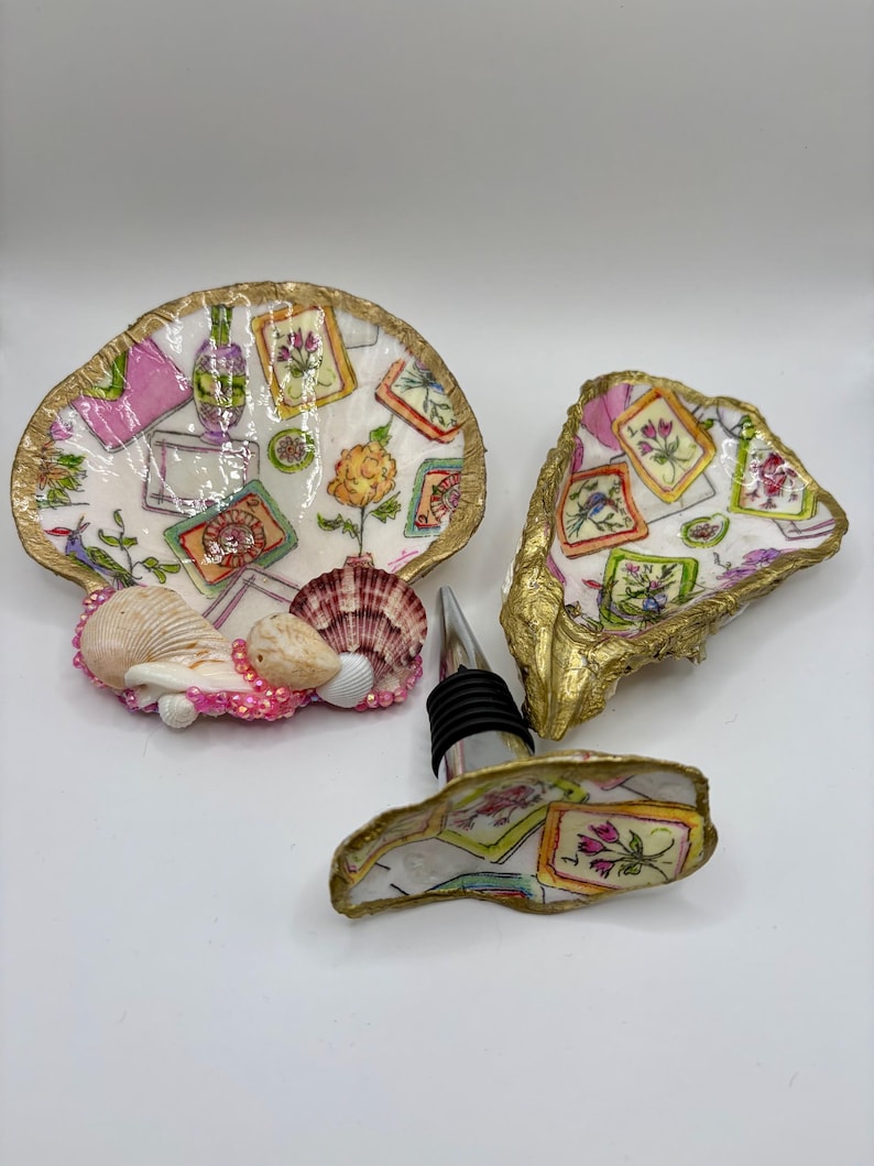 Op de afbeelding: Twee decoratieve oesterschelpen en een flessenstop met decoupage bloemen- en ingelijste kunstpatronen. De schelpen hebben goudkleurige randen en de grotere schelp is versierd met kleine roze kraaltjes en extra natuurlijke schelpen.