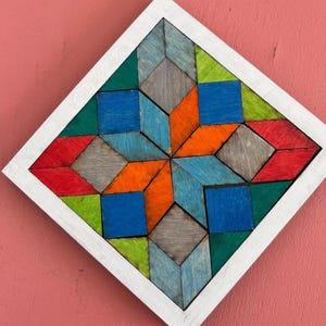 May include: A colorful wooden geometric art piece with a white frame. The artwork features a starburst design composed of various geometric shapes in shades of blue, orange, green, red, and gray. The piece is mounted on a pink surface.