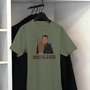 May include: Olive green t-shirt with a graphic of a person giving the three-finger salute, with the text "DREI GLÄSER!". The shirt is hanging in a white closet with other clothing items.