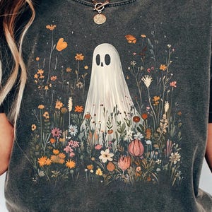 May include: Dark grey t-shirt with a whimsical illustration of a ghost in a field of colourful wildflowers and butterflies. The ghost has a simple, friendly design. The shirt has a vintage, washed look.