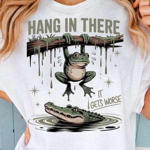 Puede incluir: Camiseta blanca con una rana de dibujos animados colgando de una rama, el texto de arriba dice "HANG IN THERE". Debajo, un caimán nada en el agua, con el texto "IT GETS WORSE".