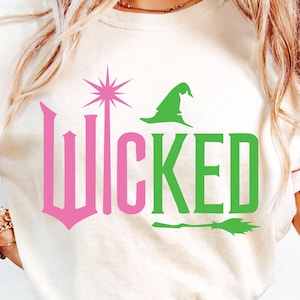 May include: A cream-coloured t-shirt featuring the word "Wicked" in pink and green. The design includes a pink starburst, a green witch's hat, and a broom. A casual, everyday wear item.