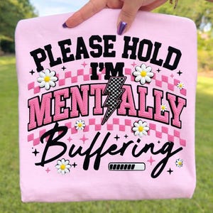 May include: Pink sweatshirt with the text "PLEASE HOLD I'M MENTALLY BUFFERING" in black and pink lettering. The design includes a lightning bolt, daisies, and a loading bar graphic. The sweatshirt is held up by a hand.
