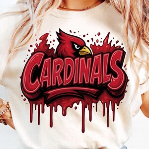 May include: A cream-coloured t-shirt featuring a graphic of a red cardinal bird head above the word CARDINALS in bold red lettering. The design includes a dripping paint effect in shades of red, creating a textured, layered appearance across the chest.