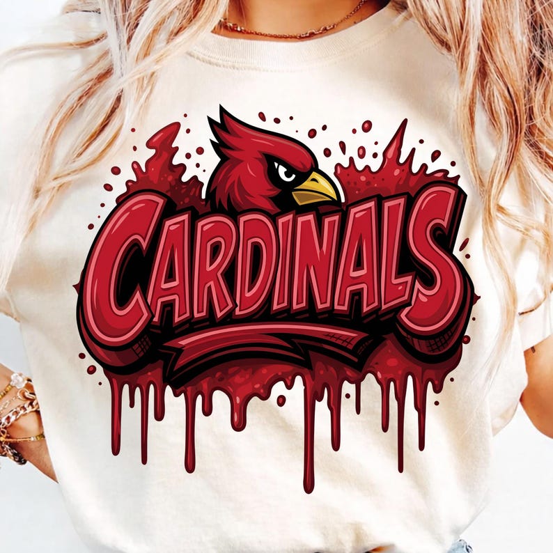 May include: A cream-coloured t-shirt featuring a graphic of a red cardinal bird head above the word CARDINALS in bold red lettering. The design includes a dripping paint effect in shades of red, creating a textured, layered appearance across the chest.