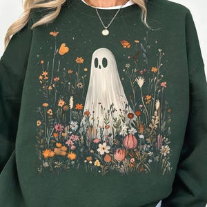 May include: Dark green sweatshirt featuring a whimsical illustration of a ghost in a field of colorful wildflowers and butterflies. The ghost has a simple, minimalist design. The flowers are in shades of orange, pink, and white.