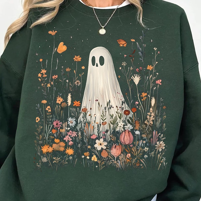 May include: Dark green sweatshirt featuring a whimsical illustration of a ghost in a field of colorful wildflowers and butterflies. The ghost has a simple, minimalist design. The flowers are in shades of orange, pink, and white.