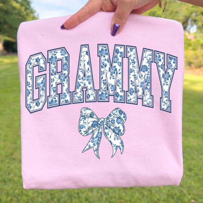 May include: A light pink sweatshirt with the word "GRAMMY" in navy blue block letters filled with a blue floral pattern. A matching blue floral bow is below the word.