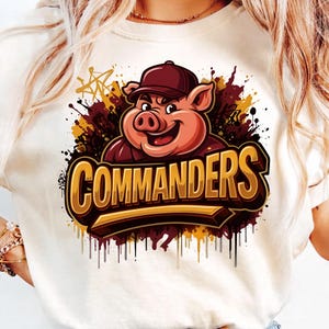 May include: A cream-coloured t-shirt with a cartoon pig wearing a maroon baseball cap. The pig is smiling, with the word "COMMANDERS" in gold and brown lettering below. The design includes paint splatter effects.