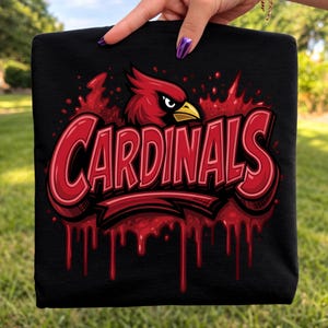 May include: A black t-shirt featuring a graphic of a red cardinal bird head above the word Cardinals in bold red lettering. The design includes a red paint splatter effect with drips extending downward from the text, creating a layered appearance.