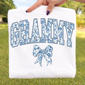 May include: White sweatshirt with the word "GRAMMY" in navy blue block letters filled with a blue floral pattern. A matching blue floral bow is below the word. The sweatshirt is held up by a hand.