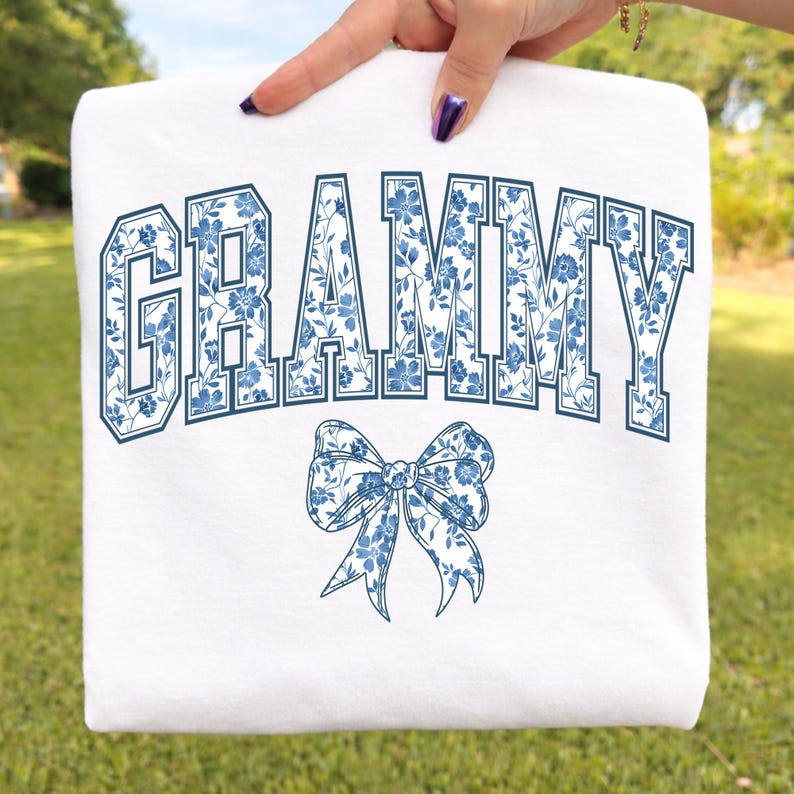 May include: White sweatshirt with the word "GRAMMY" in navy blue block letters filled with a blue floral pattern. A matching blue floral bow is below the word. The sweatshirt is held up by a hand.