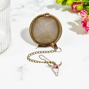 May include: A vintage-style tea infuser with a round, mesh basket and a decorative chain. The chain ends with a charm shaped like a bull skull, adorned with colorful floral accents. The infuser is made of brass-toned metal.